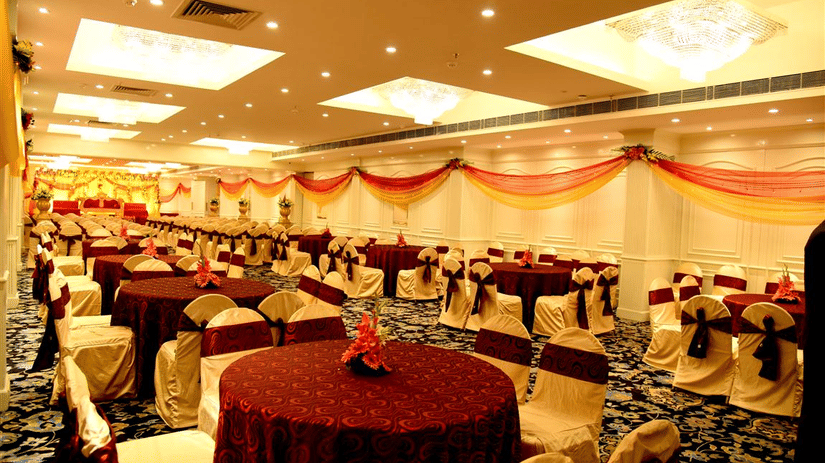 A spacious, well-lit banquet hall with red and gold decor, round tables with red tablecloths, and chairs arranged for a large event - Clarks Avadh, Lucknow