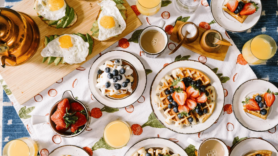 Overhead view of a breakfast spread with waffles topped with strawberries and blueberries, avocado toasts with fried eggs, orange juice, and coffee.