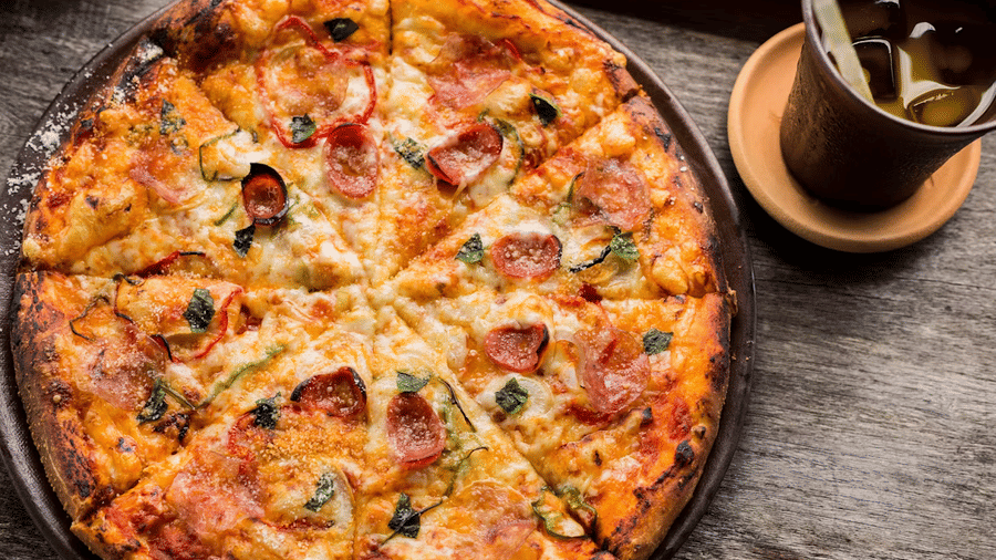 Sliced thin-crust pizza topped with salami, red pepper strips, basil, and grated cheese, served with an iced drink in a clay tumbler.