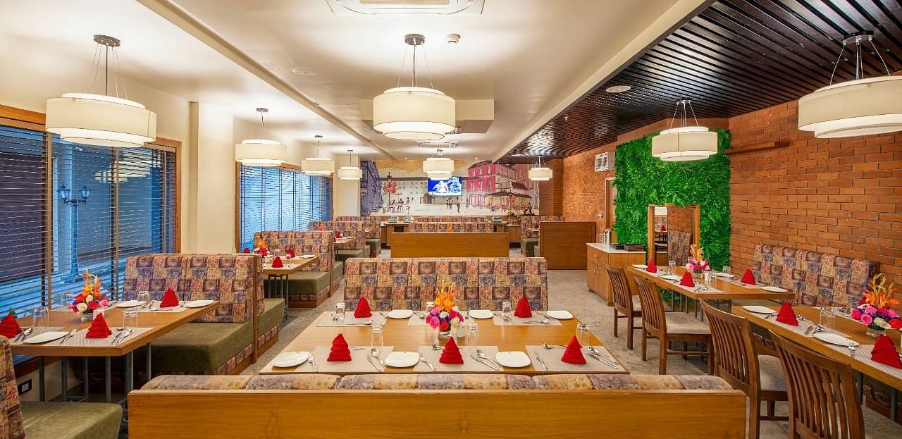 A warmly lit and inviting restaurant dining area with numerous tables set for meals, featuring patterned booth seating and wooden chairs, red napkins, and floral centrepieces, under soft pendant lighting - Lords Inn Motikhavdi, Jamnagar.