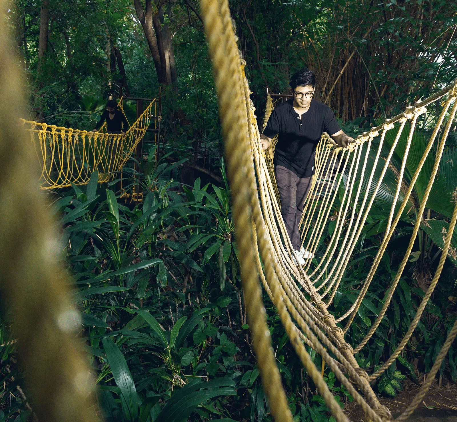A guy climbing a rope bridge that is suspended between trees, surrounded by greenery and nature near Avana Resort