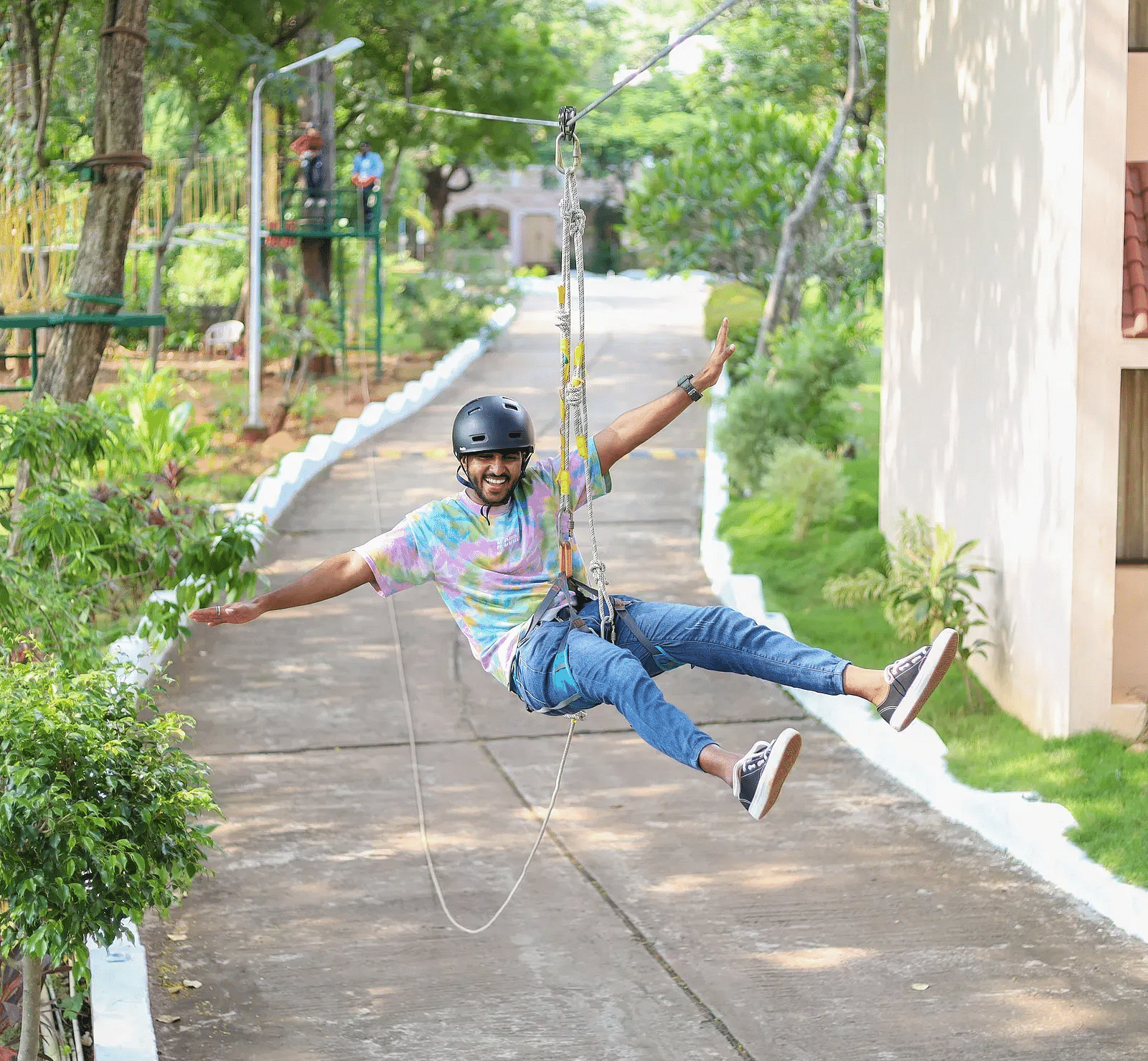Man enjoying a zipline ride through a green resort area, smiling with arms outstretched, wearing a helmet and casual clothes.