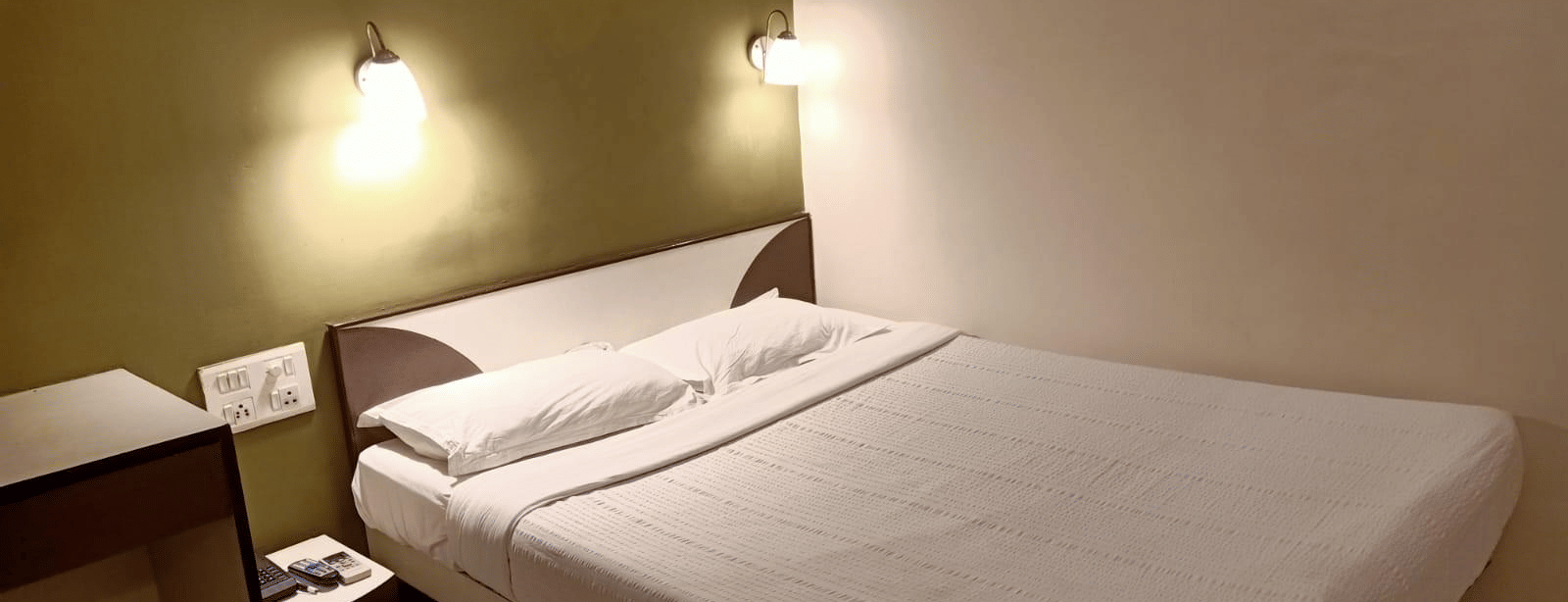 A Standard AC Room at Hotel Pandian, featuring a neatly-made bed with white sheets, 2 pillows, 2 wall lamps, and a nightstand.