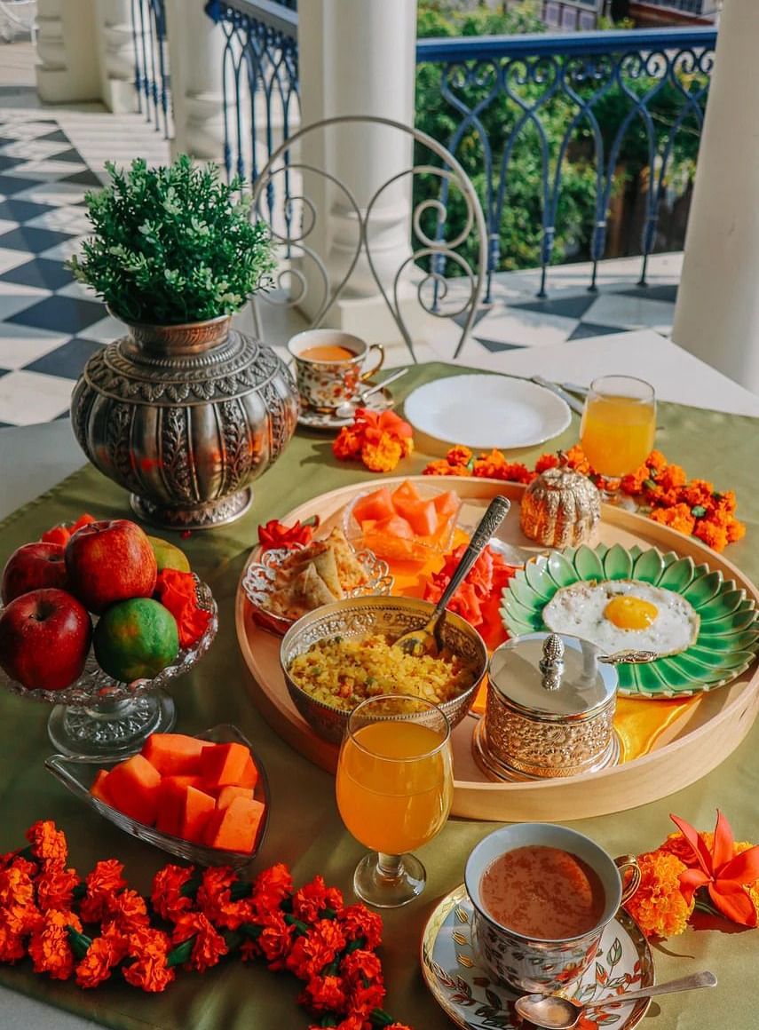A breakfast spread laid out on a table on a balcony, featuring fruits, eggs, drinks, and other dishes, with a view of blue railings and greenery | Dileep Kothi, Jaipur
