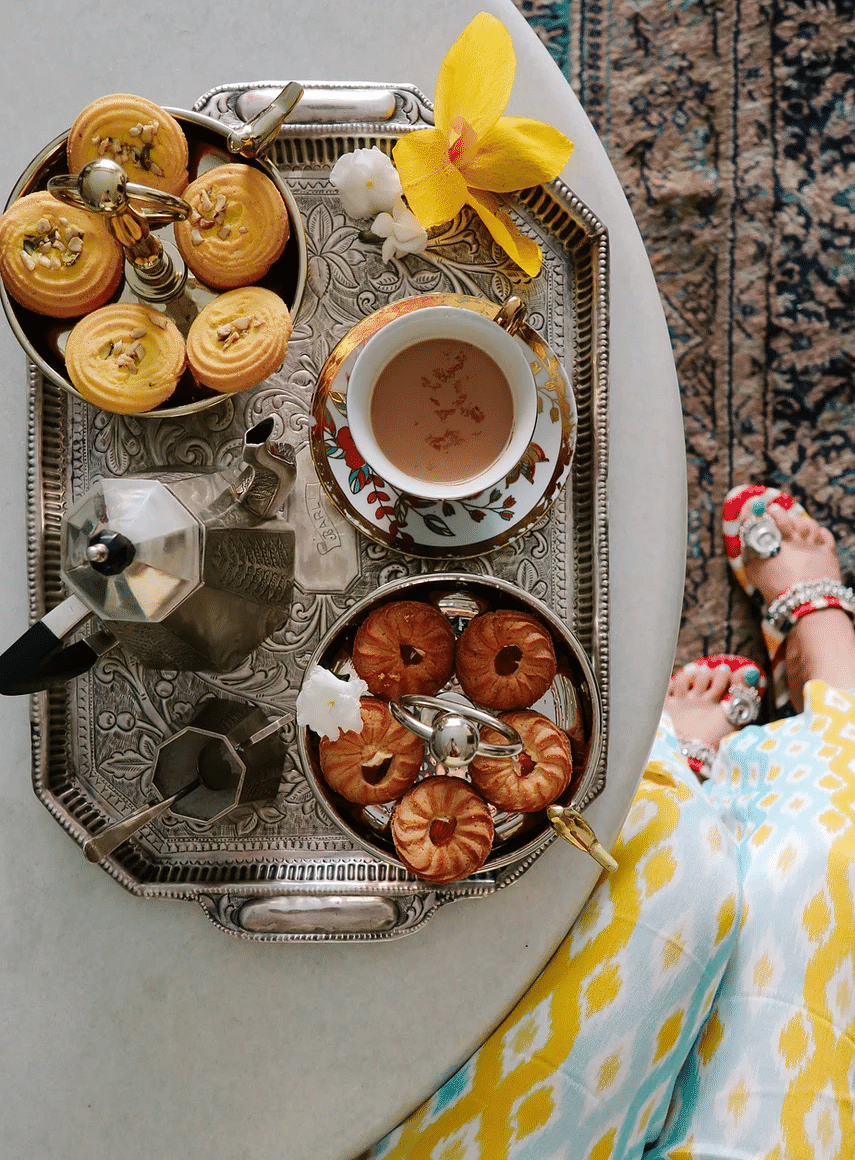A flat lay of a silver tray with coffee, pastries, and a yellow flower on a white table, with a person's patterned dress and feet visible at the edge | Dileep Kothi, Jaipur