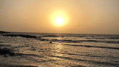 A view of the sun setting over the sea with waves reflecting the light and rocks visible in the foreground.
