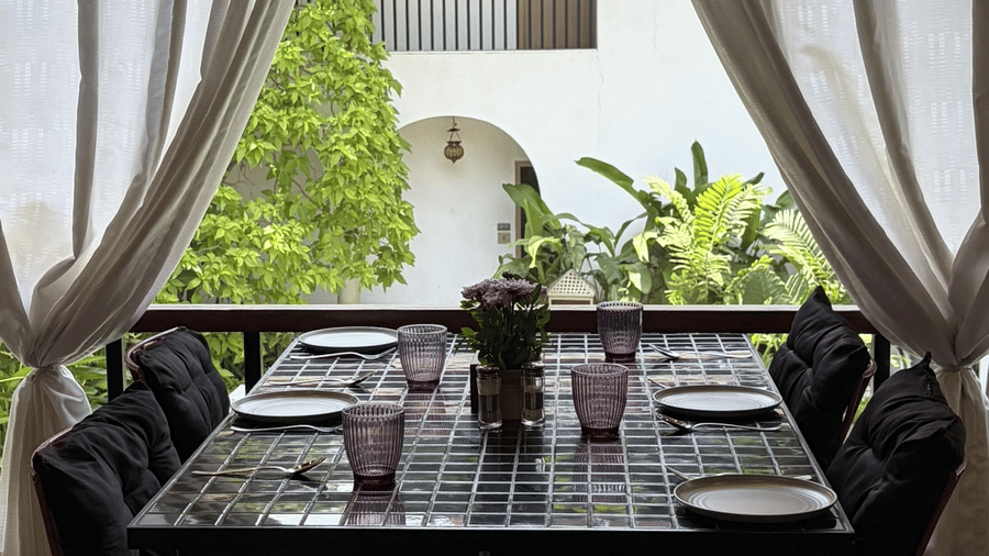 A dining table sits framed by white curtains on a balcony looking out towards a white villa at Bougainvillea, Alibaug.