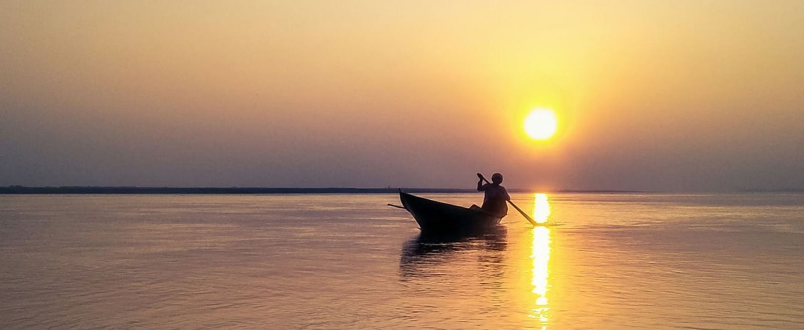 A boat with a person silhouetted against a setting sun over a calm body of water - Rhino and River BluSalzz Collection, Pobitora