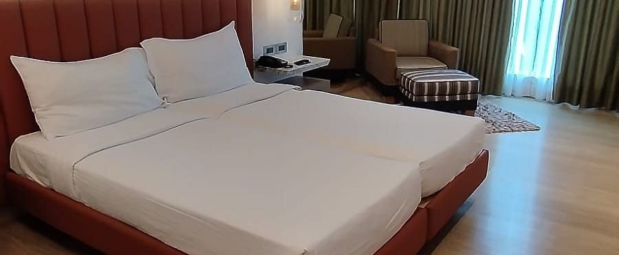 A well-lit, wooden floored, executive room in Mirage Hotel, featuring twin beds with decorative head-rest and a wall to wall window with partially closed drapes. 