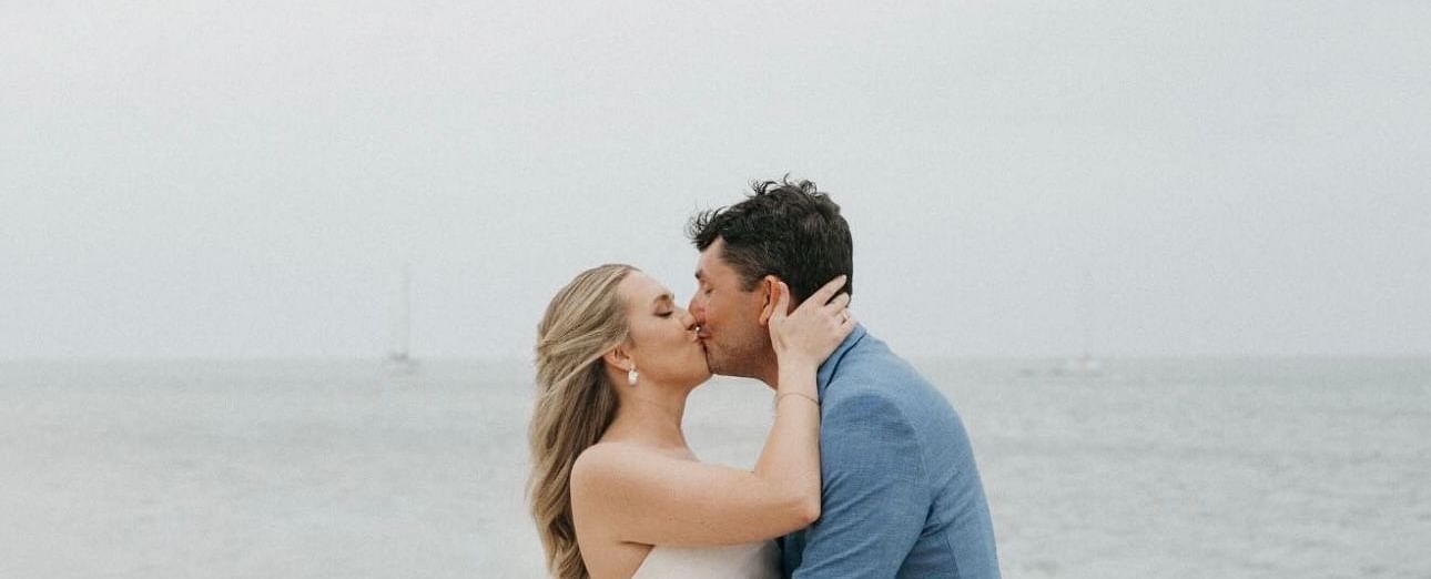 Bride and groom kissing each other in front of the ocean with a soft, cloudy sky as the backdrop.