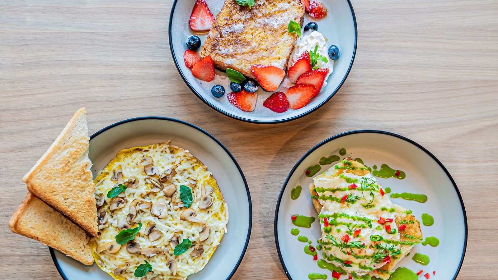 Three plates at Indie Stays Mumbai International Airport: one with mushroom omelette and basil leaves, one with toast topped with strawberries and blueberries, and one with egg toast and veggies.