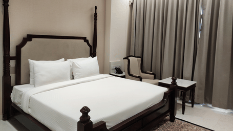A bedroom at Anant Mahal, Jaipur featuring a king size bed with multiple pillows arranged at the head of the bed, a patterned rug, floor to ceiling curtains behind a table, and a chair.