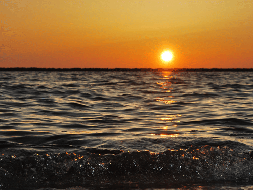A vibrant orange and yellow sunset over the sea is reflected on the wet sand of a beach with gentle waves.