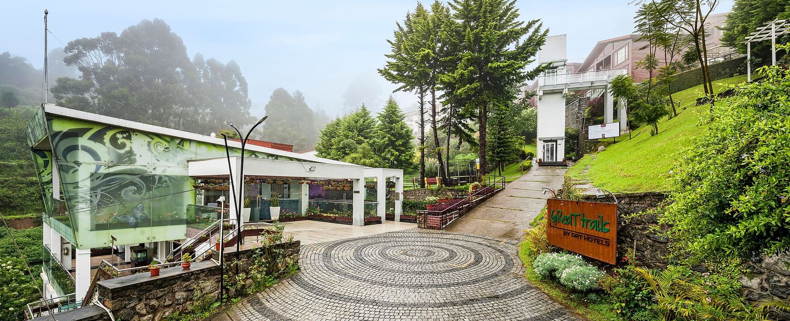 A facade image of the GReaT Trails Kodaikanal By GRT Hotels with lush greenery and the trails leading to the best mountain view resort, Kodaikanal, Tamil Nadu