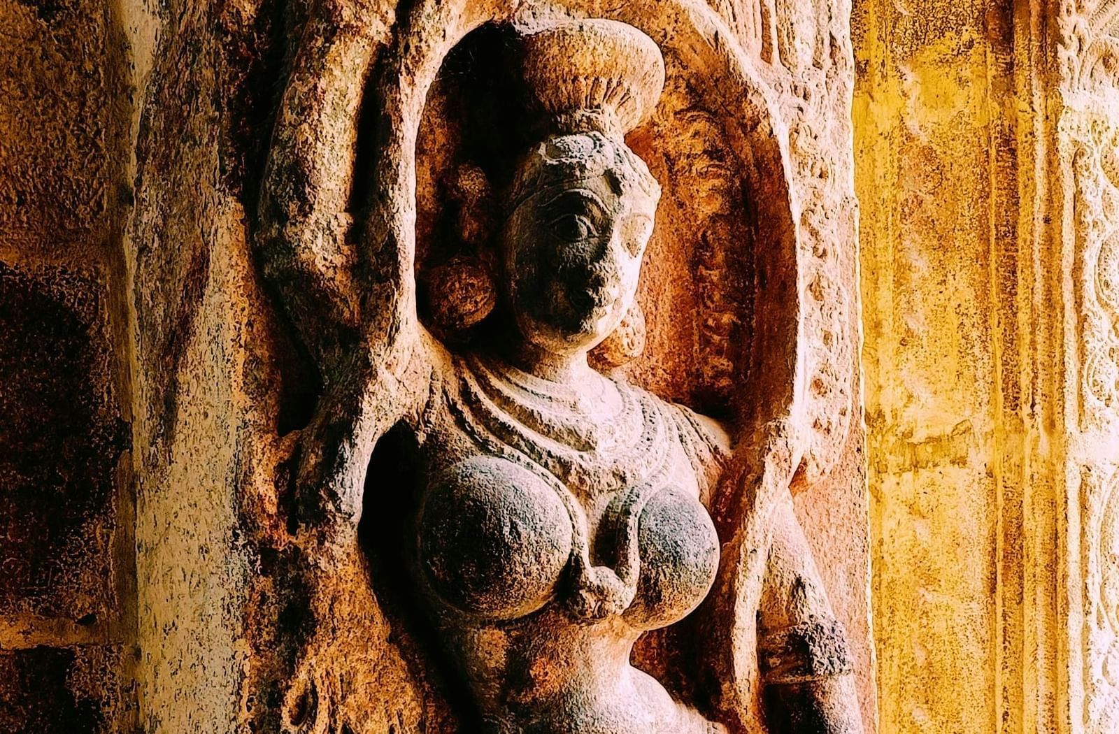  A vertical close-up of a detailed stone carving on an ancient temple column, depicting a female deity in an intricate, sensual pose, bathed in warm, golden light.