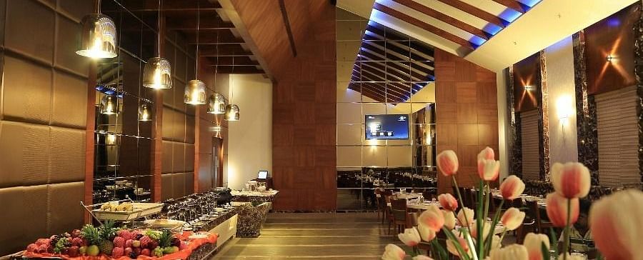 A view of the Aroma restaurant in Dhanalakshmi Srinivasan Hotels, Perambalur, featuring a buffet counter on the left and dining tables in the background. The restaurant has wooden ceiling beams and a floral arrangement in the foreground.