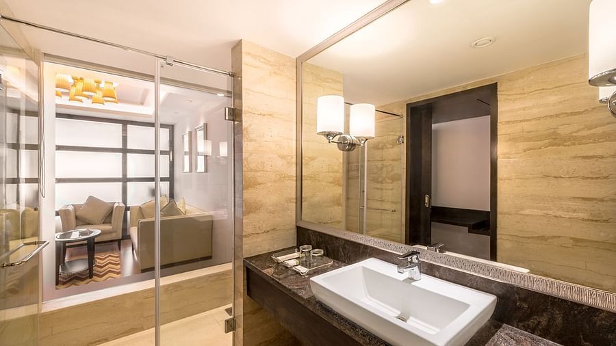 A bathroom with glass shower, large mirror and view into living area of the hotel room - TGI Grand Fortuna, Hosur