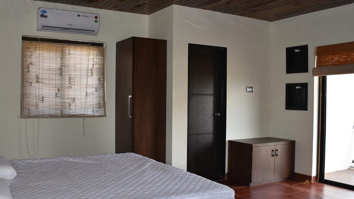 A bedroom with a white bed, dark wood furniture, a window with patterned curtains, and a sliding door leading to a balcony.