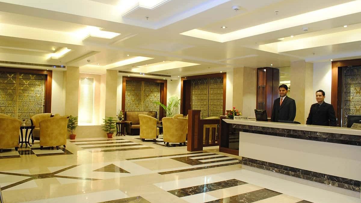 A hotel lobby with a reception desk staffed by two individuals, seating areas, and patterned flooring.