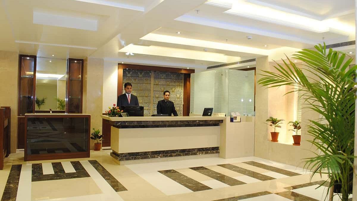 A brightly lit hotel reception area with two staff members behind a counter, patterned flooring, and decorative plants.