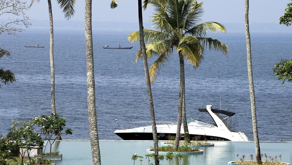 An image of a boat gently floats on a lake surrounded by coconut trees - Rhythm Kumarakom