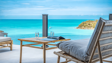 An outdoor patio with comfortable seating and a table overlooking a vibrant blue ocean under a sunny sky.
