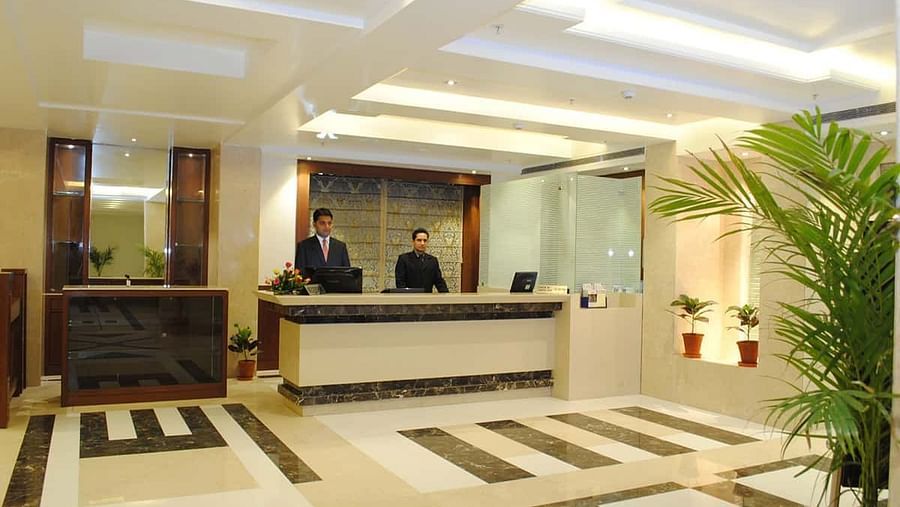 A brightly lit hotel reception area with two staff members behind a counter, patterned flooring, and decorative plants.