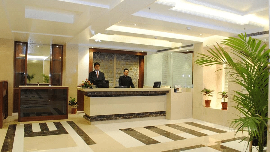 A brightly lit hotel reception area with two staff members behind a counter, with patterned flooring, and decorative plants at Libra Lords Inn Jaipur