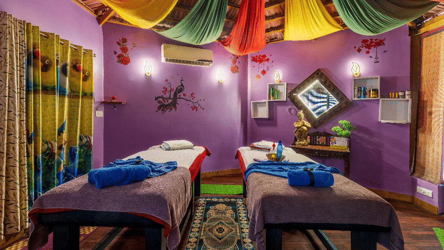 A purple spa room featuring 2 massage beds with blue towels, colourful draped fabric on the ceiling, and rugs at Amantra Shilpi Resort, Udaipur.