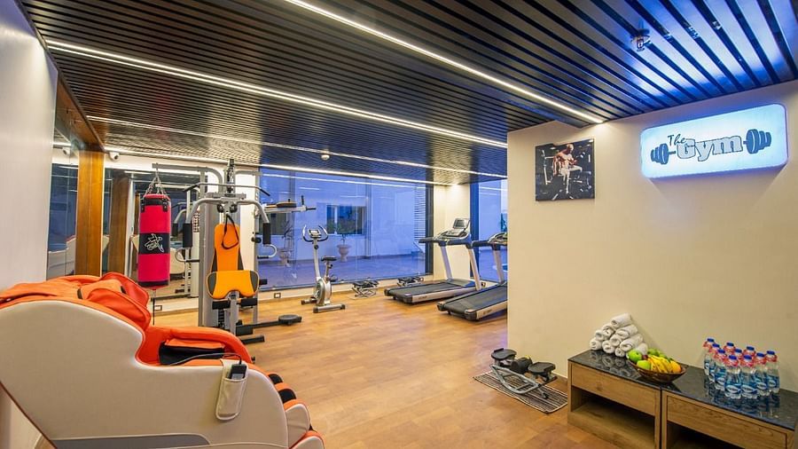 A modern gym area featuring various exercise equipment including treadmills, weight machines, a red and black punching bag, and a massage chair, with a wooden floor and a ceiling with strip lighting and a blue illuminated sign - Lords Inn Motikhavdi, Jamna