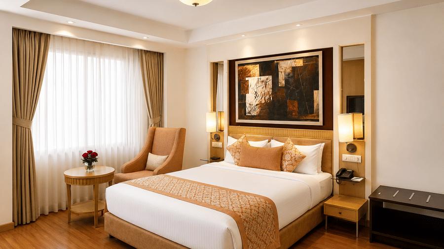 Hotel room with a bed dressed in white and gold linen, a beige armchair, a wall-mounted artwork and long sheer curtains by the window nearby.