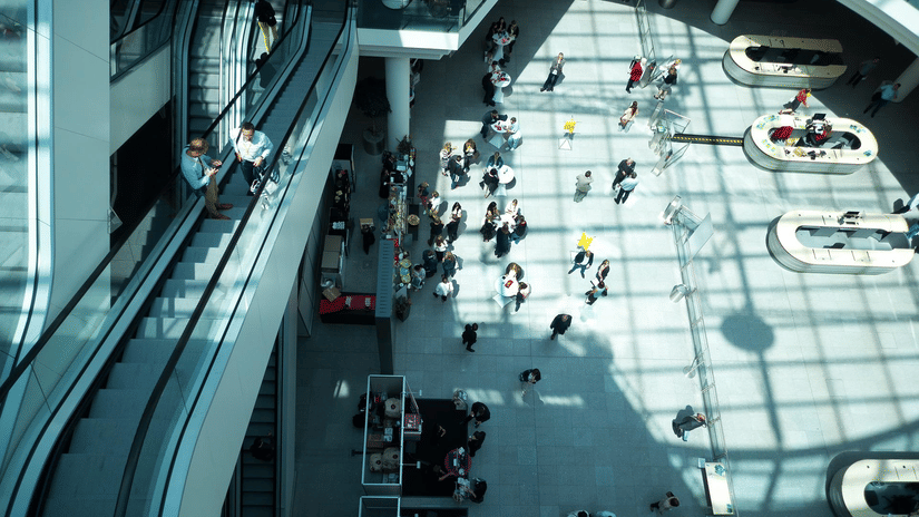 A top-down view of a convention centre with an escalator on the left hand side, and people near the registration desk and other showing areas at ITB India 2026.