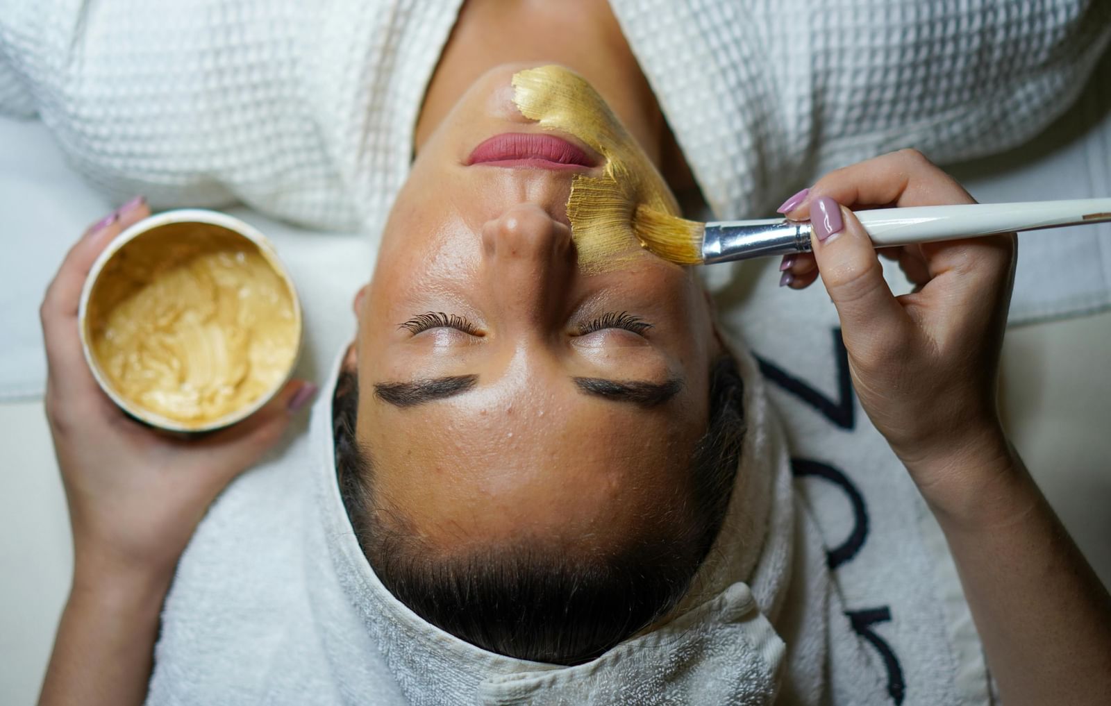 A woman relaxes as a beautician applies a golden facial mask with a soft brush during a spa treatment.