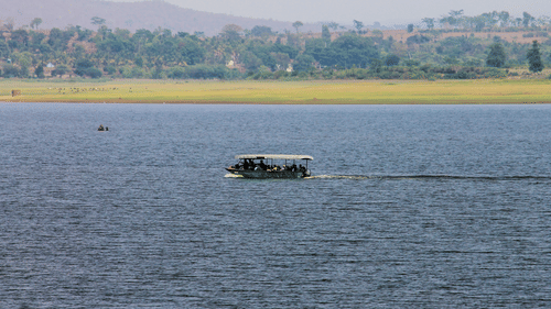 A river scene with a motorised boat carrying passengers across calm blue waters. The far shore has a grassy riverbank with scattered trees and hills in the background under a hazy sky.