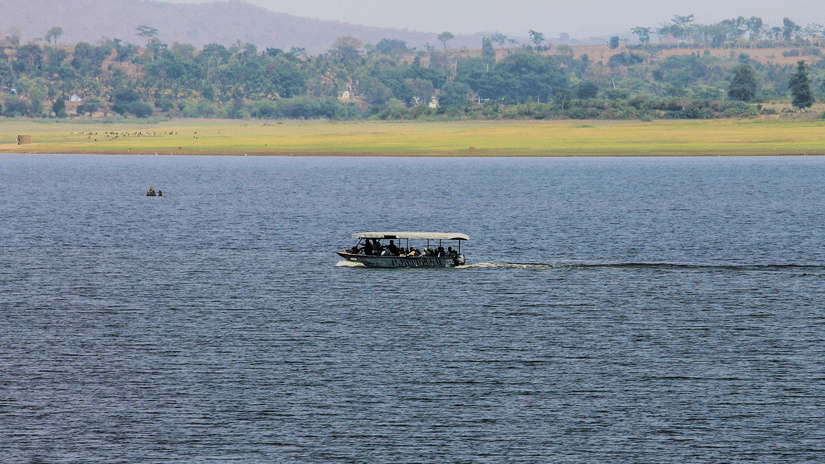 A river scene with a motorised boat carrying passengers across calm blue waters. The far shore has a grassy riverbank with scattered trees and hills in the background under a hazy sky.