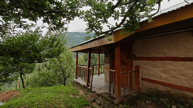 Exterior of the Deo Tibba Cottage featuring the entrance surrounded with a lush green lawn and trees, with a backdrop of the hills at Amara Upepo - The Sky Village, Manali.
