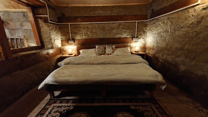 A close-up shot of a double bed with a pattered carpet in front of it at the Friendship Peak Cottage featuring a wooden ceiling above it at Amara Upepo - The Sky Village, Manali.