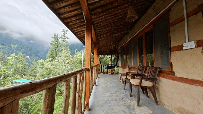 The porch area of the Deo Tibba Cottage with 2 arm chairs kept against the wall overlooking the mesmerising green forests with the backdrop of the misty hills at Amara Upepo - The Sky Village, Manali.