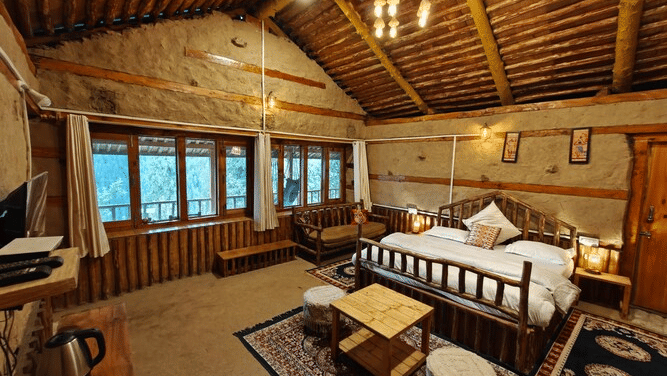 Full-view shot of the brightly-lit interiors of the Deo Tibba Cottage featuring medium-sized windows overlooking the forest outside at Amara Upepo -The Sky Village, Manali.
