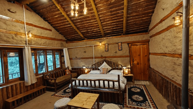 A brightly-lit interior of the Deo Tibba Cottage featuring a double bed in the middle along with a coffee table in front of it with 2 stools around it at Amara Upepo - The Sky Village, Manali.