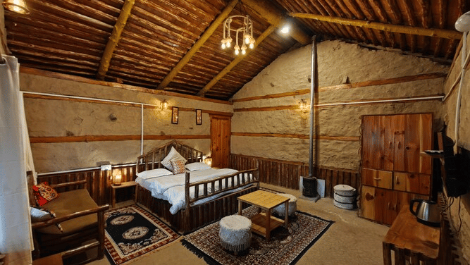 Corner view of the Deo Tibba Cottage showcasing the a double bed, a coffee table placed over a patterned carpet, and a wooden wardrobe in the corner of the room at Amara Upepo - The Sky Village, Manali.