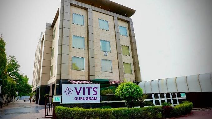A low-angle exterior shot of a multi-storey building with a sign that reads VITS GURUGRAM, surrounded by greenery