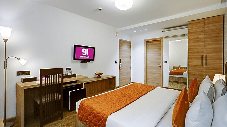A view of a guest room highlighting the wooden wardrobe, wall-mounted TV with branding, and a bed with orange accents - Hotel 91 HUDA City Centre, Gurugram