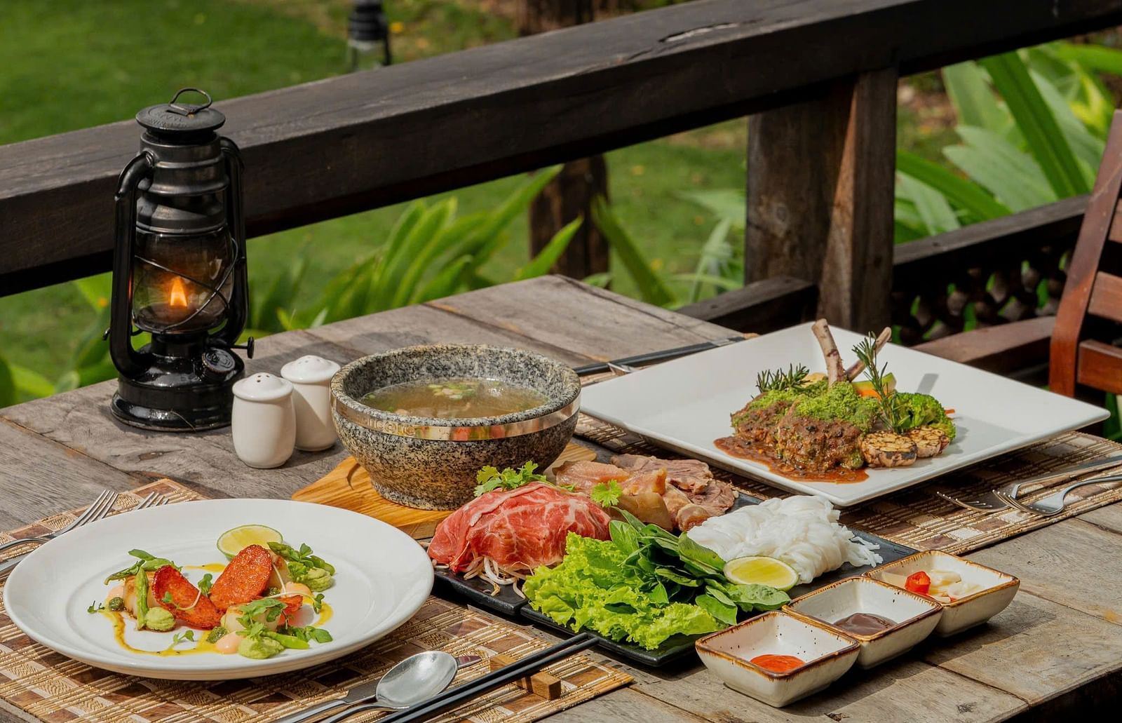 Outdoor table set with dishes, cutlery, and a lantern, on a patio with greenery.
