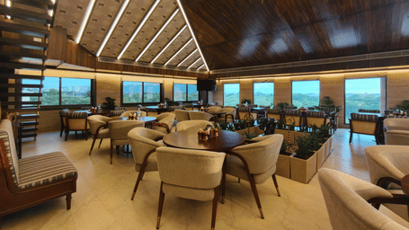 Spacious dining room at The Andhra Leaf restaurant, Ni Central Park, Mysore, featuring round dining tables, comfortable armchairs, and a slanted wooden ceiling with recessed lighting.