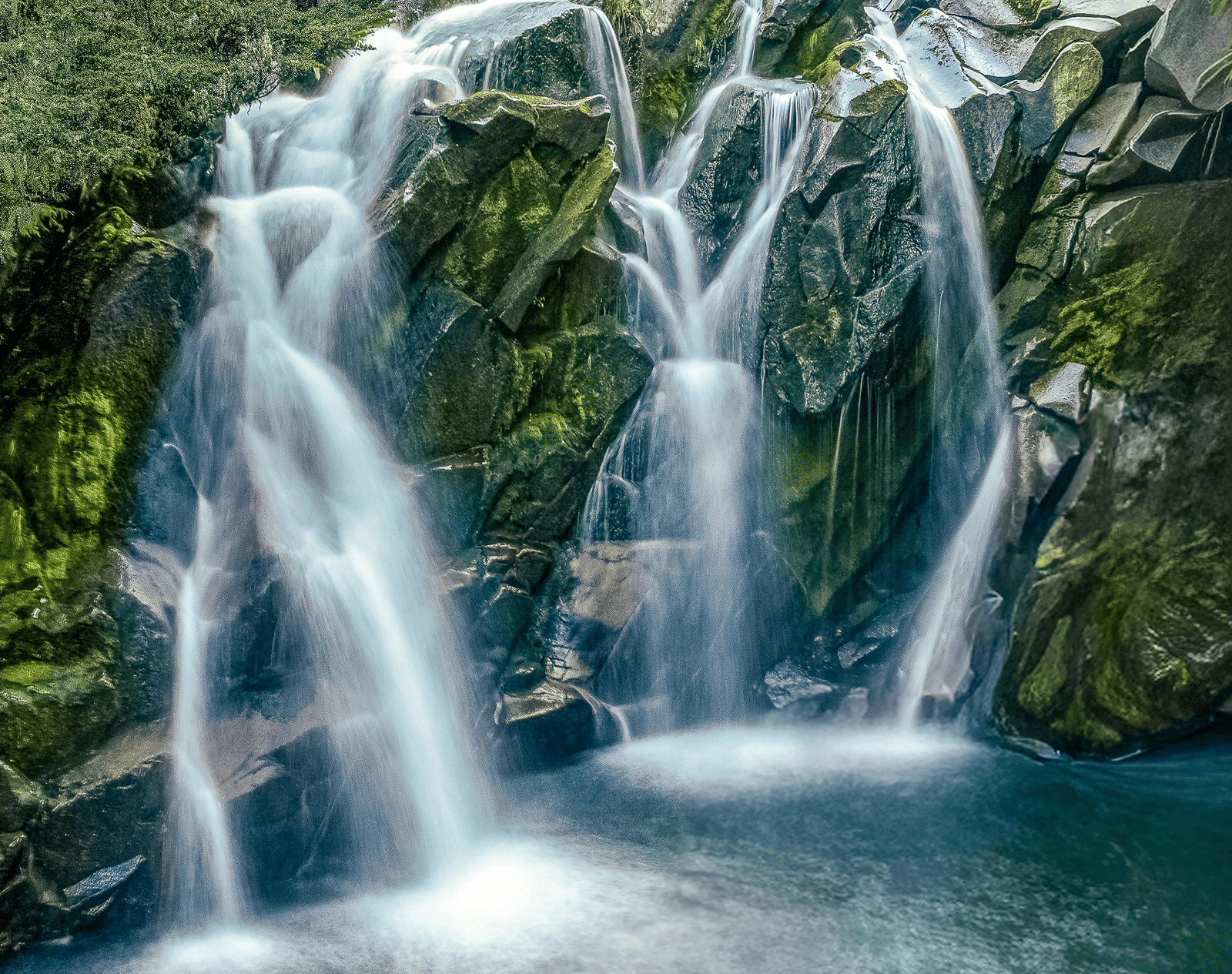 A multi-tiered waterfall cascading over rocky terrain into a pool of water, surrounded by dense green trees