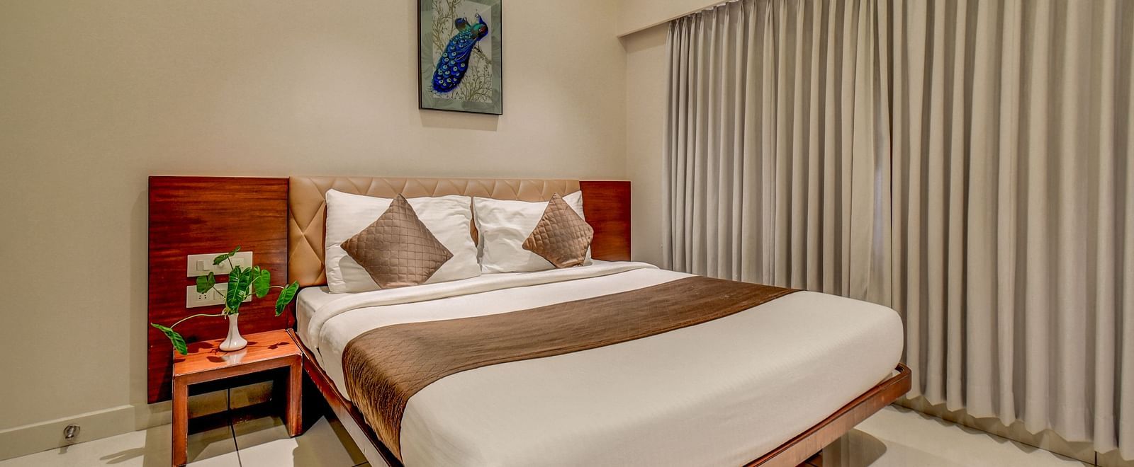 A spacious room at Harigovinda's JNStay with a large bed, a bedside table, large windows fully covered with curtains, and a wall-mounted photo frame above the headboard