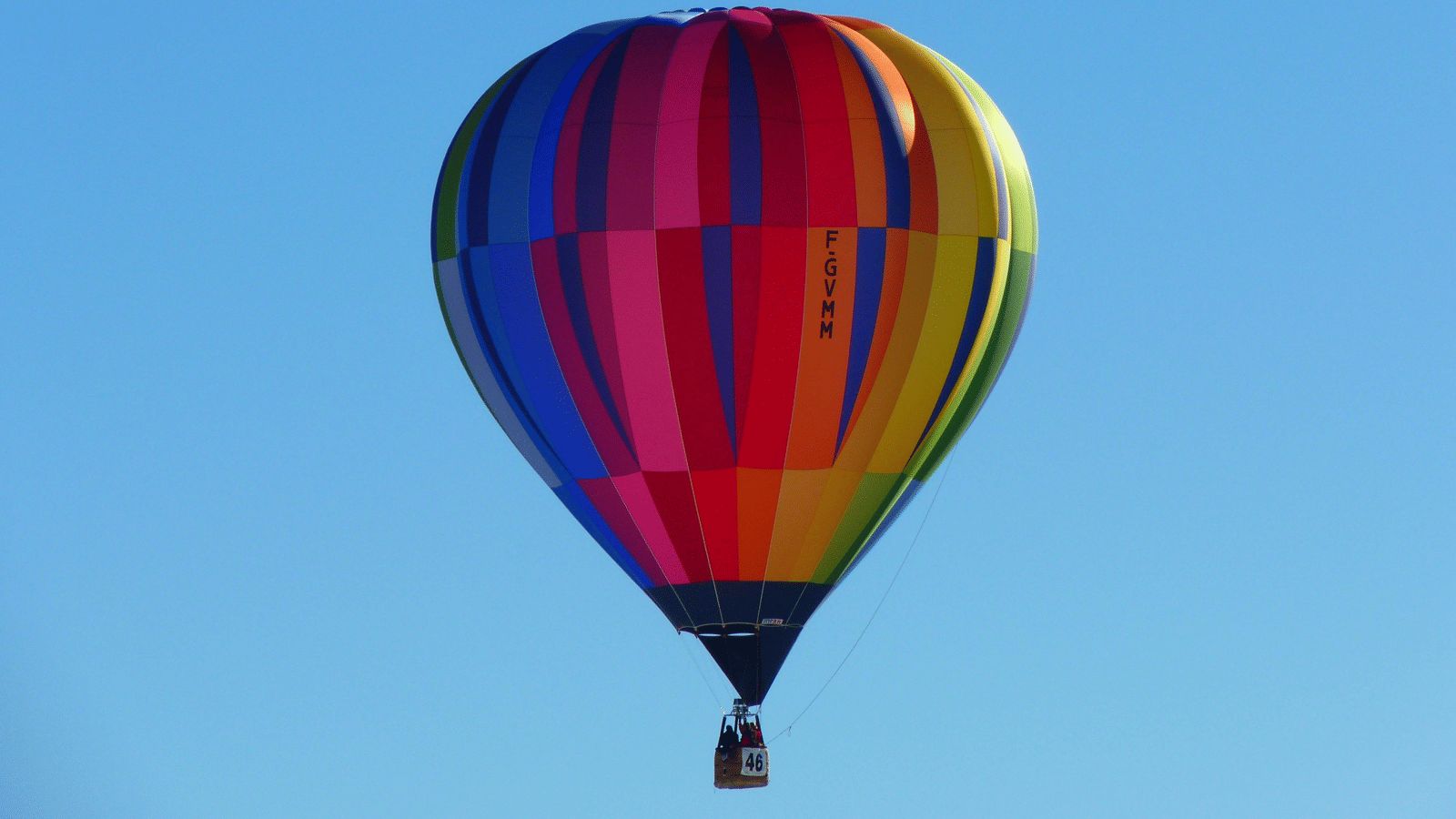 A single hot air balloon with vertical stripes in bright rainbow colours (red, pink, blue, yellow) is suspended in a clear, vivid blue sky.