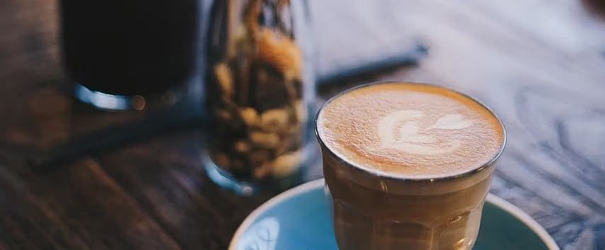 A cup of a coffee served on a blue coaster with a small vase holding dried flowers.