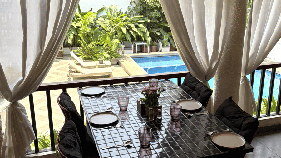An overhead view shows a dining table set on a balcony next to curtains, railings, and a swimming pool at Bougainvillea, Alibaug.